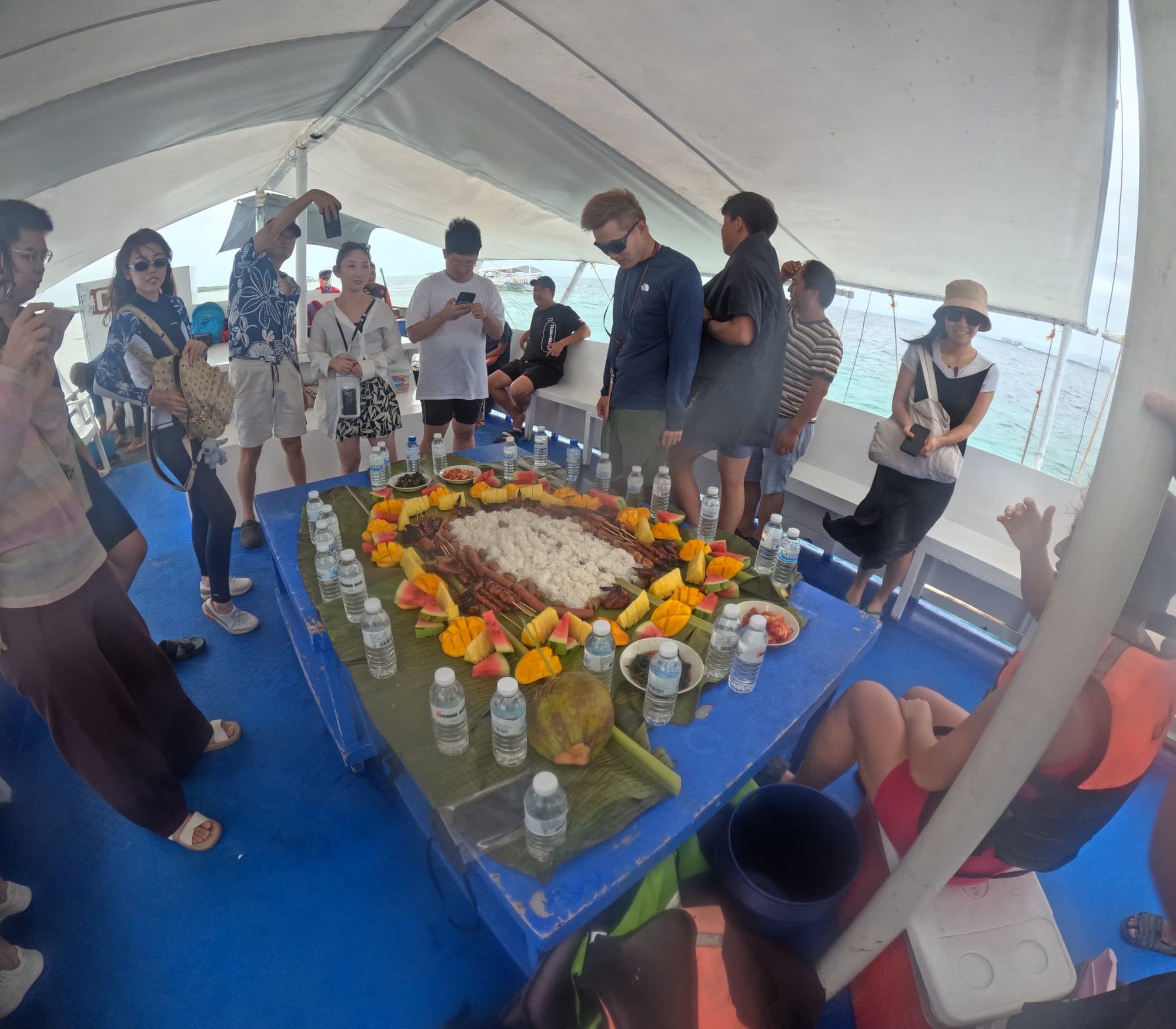 跳岛船甲板上的 boodle fight 午餐：香蕉叶铺开一长条，中间堆白米饭，两侧摆烤肉和当季水果，学员围站取餐